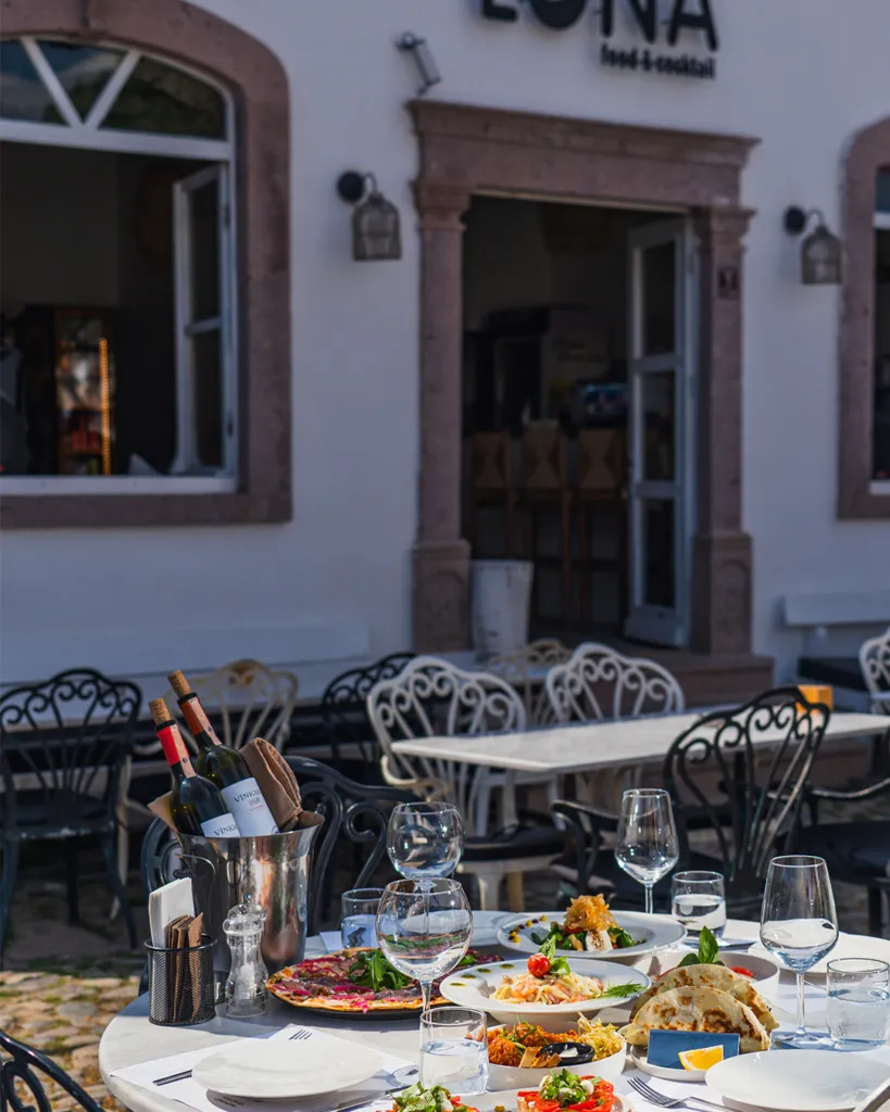 Cunda'da gezilecek yerler arasında keyifli bir durak olan LONA food & cocktail adlı restoranın dış mekanında, beyaz yuvarlak bir masanın üzerinde özenle hazırlanmış mezeler, pizzalar, makarnalar ve şarap kadehleriyle dolu şık bir sofra düzeni, arka planda ise daha fazla masa ve restoran girişi görünüyor.