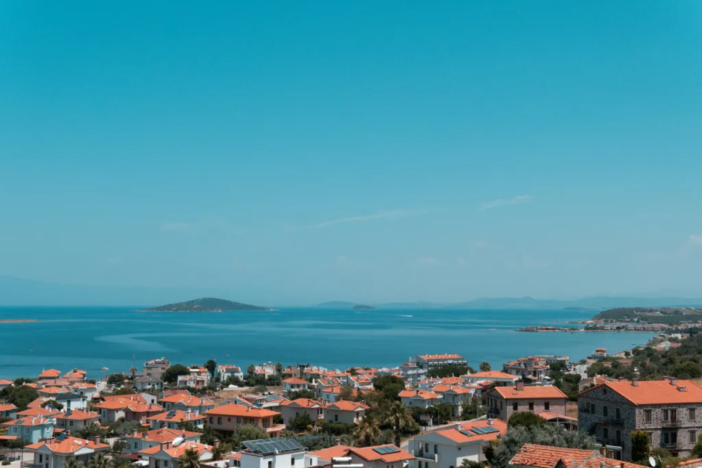 Cunda Adası'nın yukarıdan çekilmiş bu etkileyici fotoğrafında, turuncu kiremit çatılı evlerin denize doğru uzandığı, masmavi Ege Denizi'nin ve uzaktaki yeşil adacıkların belirginleştiği, buranın ne kadar güzel bir gezilecek yer olduğunu gösteren güneşli bir sahil kasabası panoraması.