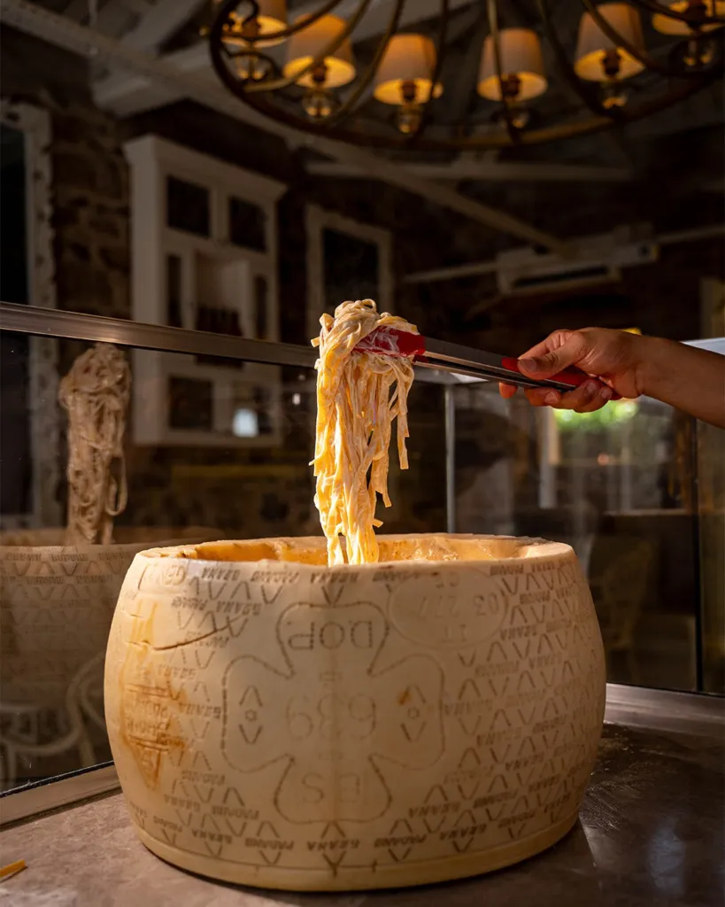 Bir elin maşayla büyük bir Grana Padano peynir tekerinin içinden kremsi makarnayı kaldırdığı bu çekim, Cunda'da gezilecek yerlerden biri olan Ola Mera restoranının özgün lezzet sunumunu gözler önüne seriyor.