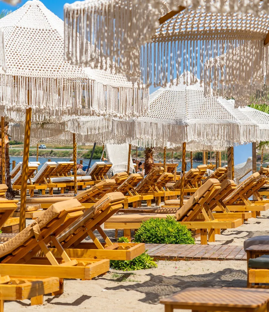 Cunda Scala Beach'te, parlak güneş altında uzanan açık renkli kumların üzerindeki ahşap şezlonglar, bej minderleriyle ve her birinin üzerinde beyaz, püsküllü makrome plaj şemsiyeleriyle sıralanmış şık bir sahil düzenini gösteriyor; uzakta masmavi deniz ve karşı kıyı görünüyor.