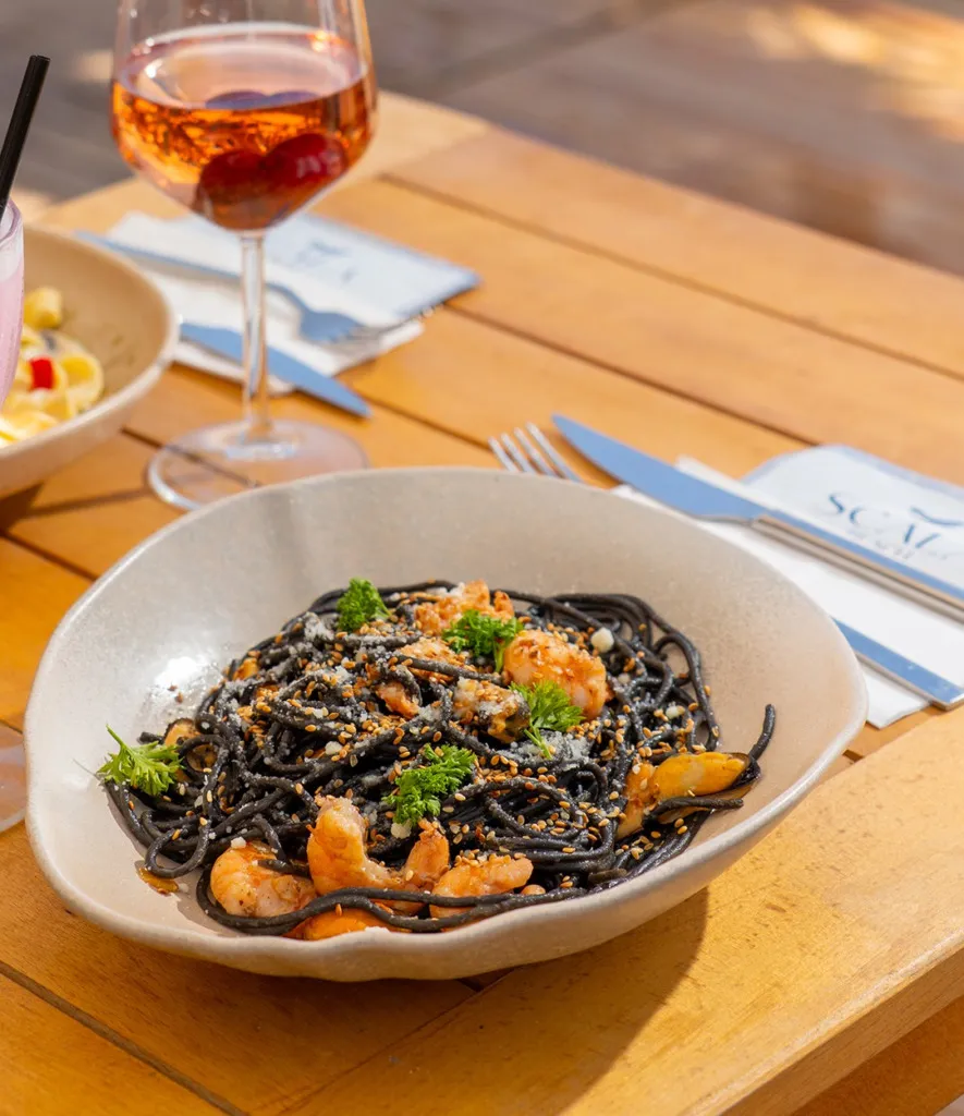Ahşap masada, Cunda'nın Scala Beach'inde güneşli bir günde servis edilen, bol karides, midye, susam ve maydanozla zenginleştirilmiş, üzerine rendelenmiş peynir serpilmiş koyu renk mürekkep balığı mürekkebi spagettisi tabağı, arka planda bir kadeh roze şarapla iştah açıcı bir görüntü sunuyor.