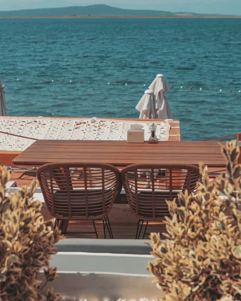 Cunda Surya Beach'teki bu karede, ön plandaki bulanık yeşil bitkilerin çevrelediği, denize bakan ahşap bir masanın üzerinde beyaz bir peçetelik ve öğütücü duruyor; masanın arkasında iki kahverengi hasır sandalye, ardından masmavi dalgalı deniz ve uzakta görünen dağlık kıyı şeridi hoş bir manzara sunuyor.
