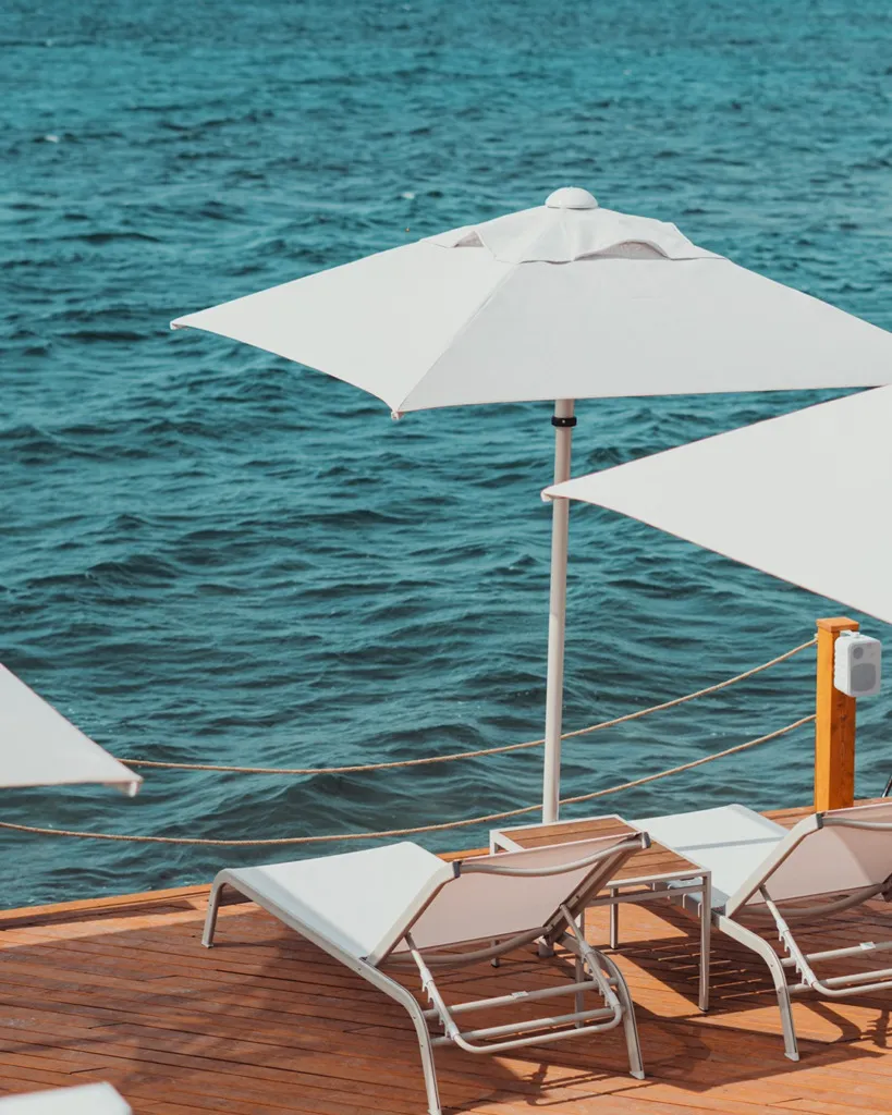 Cunda Adası'nın dingin sularına nazır ahşap iskelede, berrak turkuaz denizin kıyısında konumlanmış beyaz şemsiyeler ve konforlu şezlonglar, Ege'nin huzurlu plaj atmosferini yansıtırken, Cunda Seyahat Rehberi'mizdeki Denize Girilecek Yerler bölümünde bahsedilen Surya Beach'in zarif ve dinlendirici ortamını çağrıştırıyor.