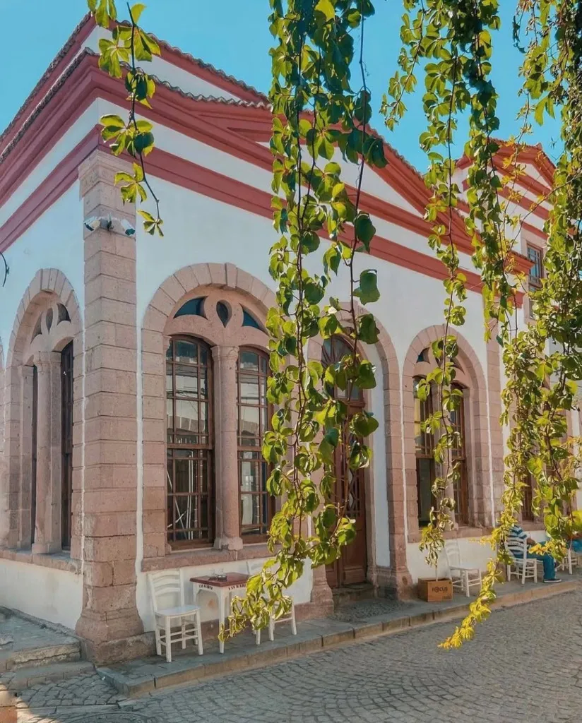 Cunda'nın Arnavut kaldırımlı sokağında, kemerli taş pencereleri ve kırmızı şeritli beyaz cephesiyle dikkat çeken tarihi Taş Kahve'nin önünde, yeşil sarmaşıkların gölgelediği masalarda oturan misafirler güneşli havanın keyfini çıkarıyor.