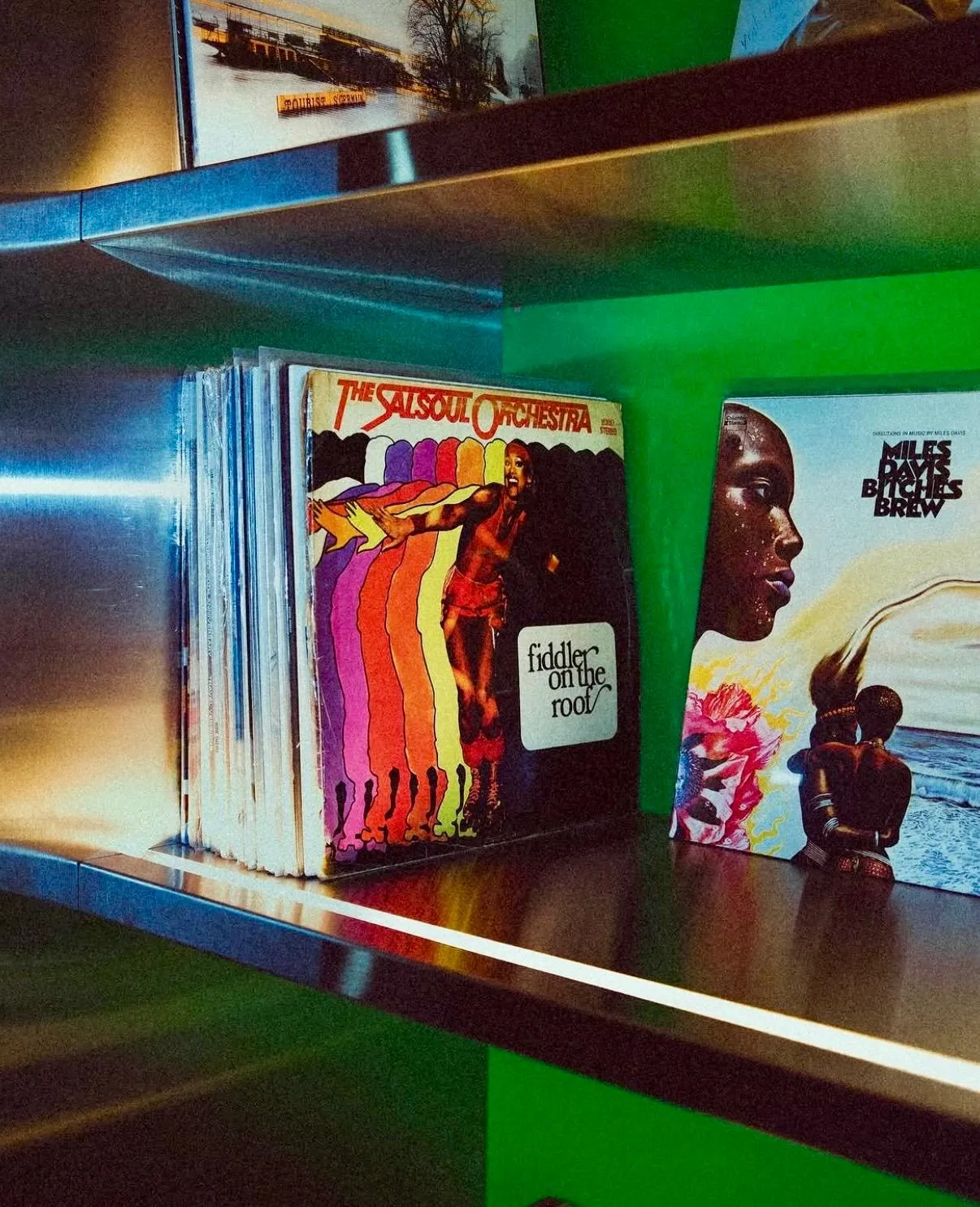 Dama Istanbul'daki parlak metal raflarda yer alan vinil plak koleksiyonundan bir kesit; ön planda Fiddler on the Roof etiketiyle The Salsoul Orchestra'nın rengarenk kapaklı albümü ve yanında Miles Davis'in Bitches Brew albümünün ikonik yüzü, yeşil duvar önünde aydınlatılarak nostaljik bir müzik deneyimi sunuyor.