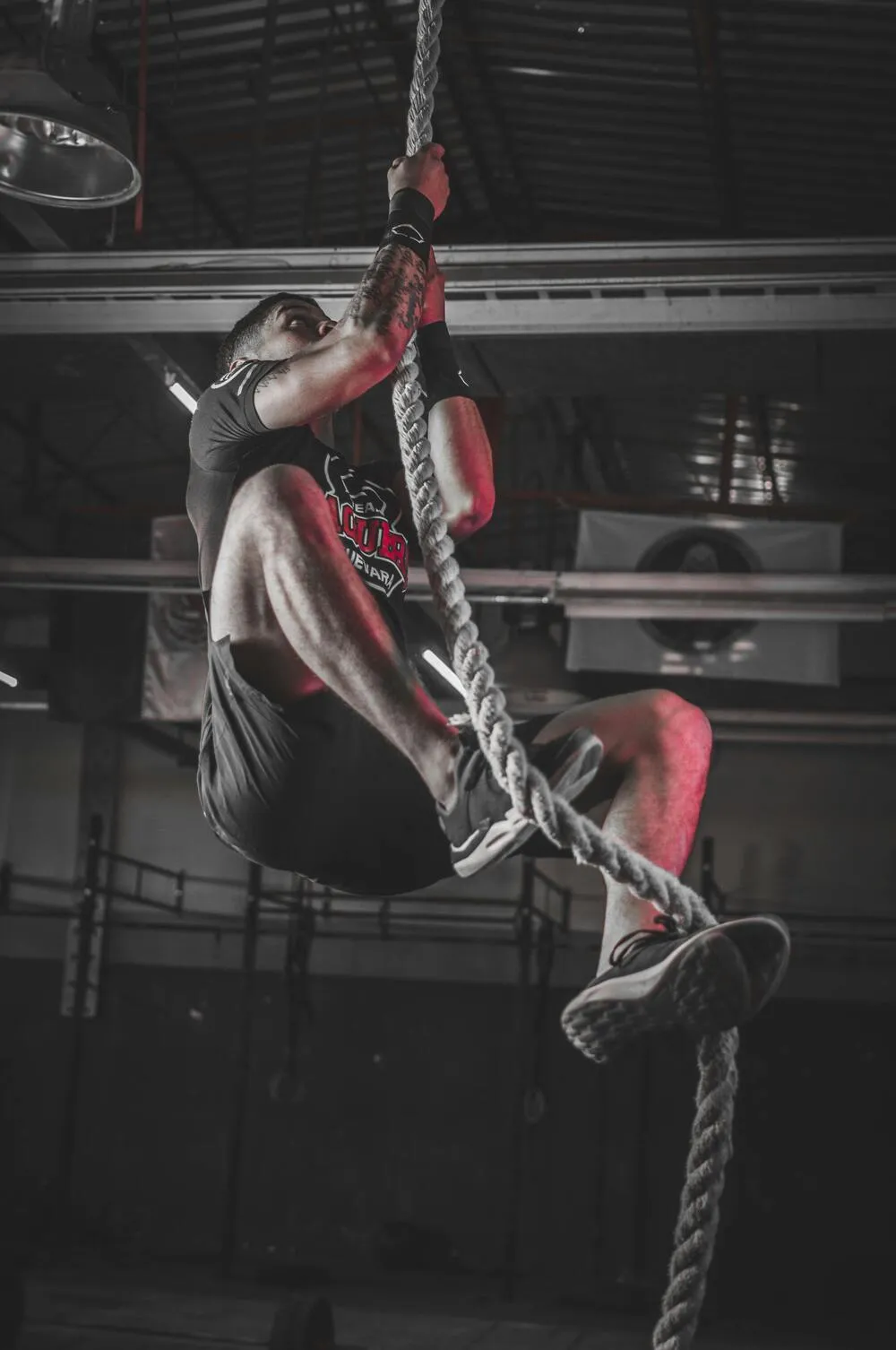 Bir fitness salonunda, dövmeli bir sporcu siyah tişört ve şortuyla, kas gücünü sergileyen zorlu bir halat tırmanışı antrenmanı yaparken, özellikle kol ve bacaklarındaki definasyon ile dikkat çekiyor.