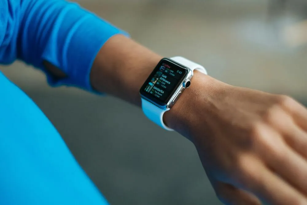 Mavi spor tişörtü giyen bir kişinin bileğinde, spor ve definasyon süreçlerini desteklemek amacıyla yakındaki kafe ve çay salonlarını (örneğin 1.4 km mesafedeki bir kafe) mesafeleriyle gösteren, saati de belirten beyaz kayışlı akıllı saat.