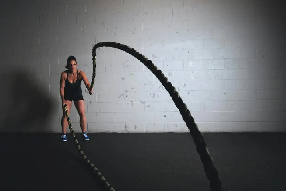 Siyah spor kıyafetleri giymiş kaslı bir kadın, loş ışıklı bir spor salonunda iki kalın savaş ipini dinamik bir şekilde kullanarak kollarını ve tüm vücudunu çalıştırarak definasyon antrenmanı yapıyor.