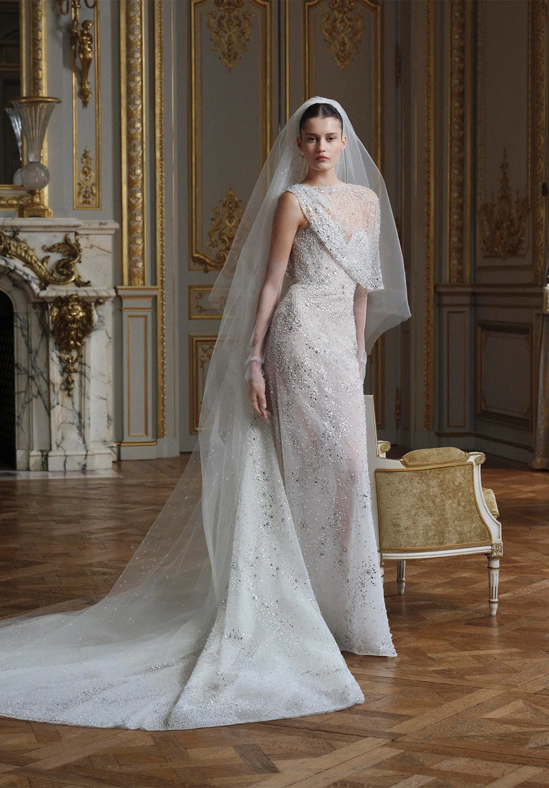 Elie Saab'ın 2026 İlkbahar koleksiyonundan, göğüs ve omuzları saran zarif şeffaf işlemeli panelleri ve drapeli yaka detayıyla öne çıkan, baştan aşağı ışıltılı kristal işlemelerle süslenmiş, uzun kuyruklu tül duvaklı couture gelinlik giyen bir model, lüks altın varaklı bir salonda duruyor.