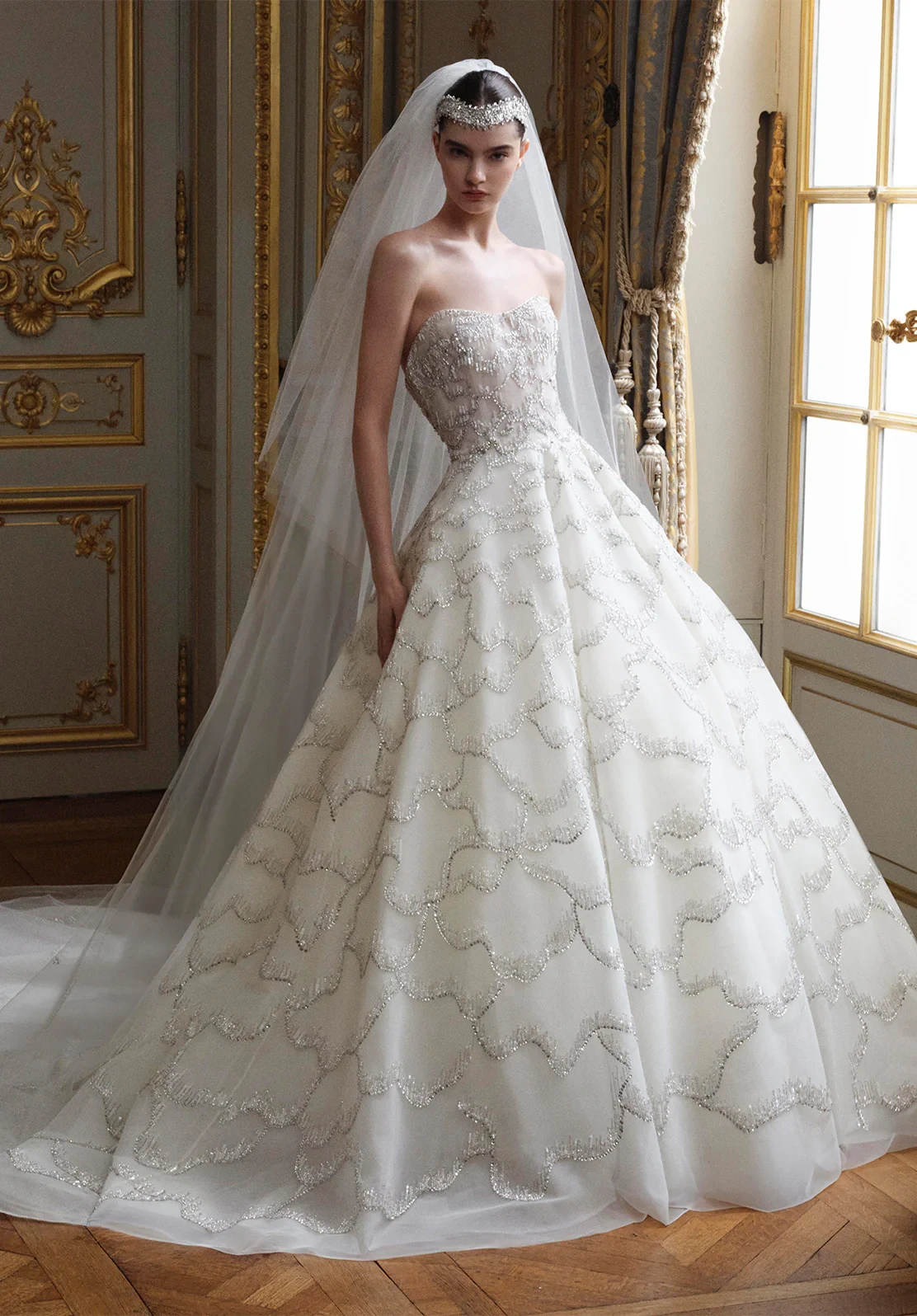 Elie Saab gelinlik koleksiyonundan, işlemeli straplez korsajı ve ışıltılı dalgalı süslemelerle bezeli hacimli eteğiyle dikkat çeken, lüks ve zarif bir gelinlik modeli, uzun tül duvak ve kristal taçla kraliyetvari bir duruş sergiliyor.