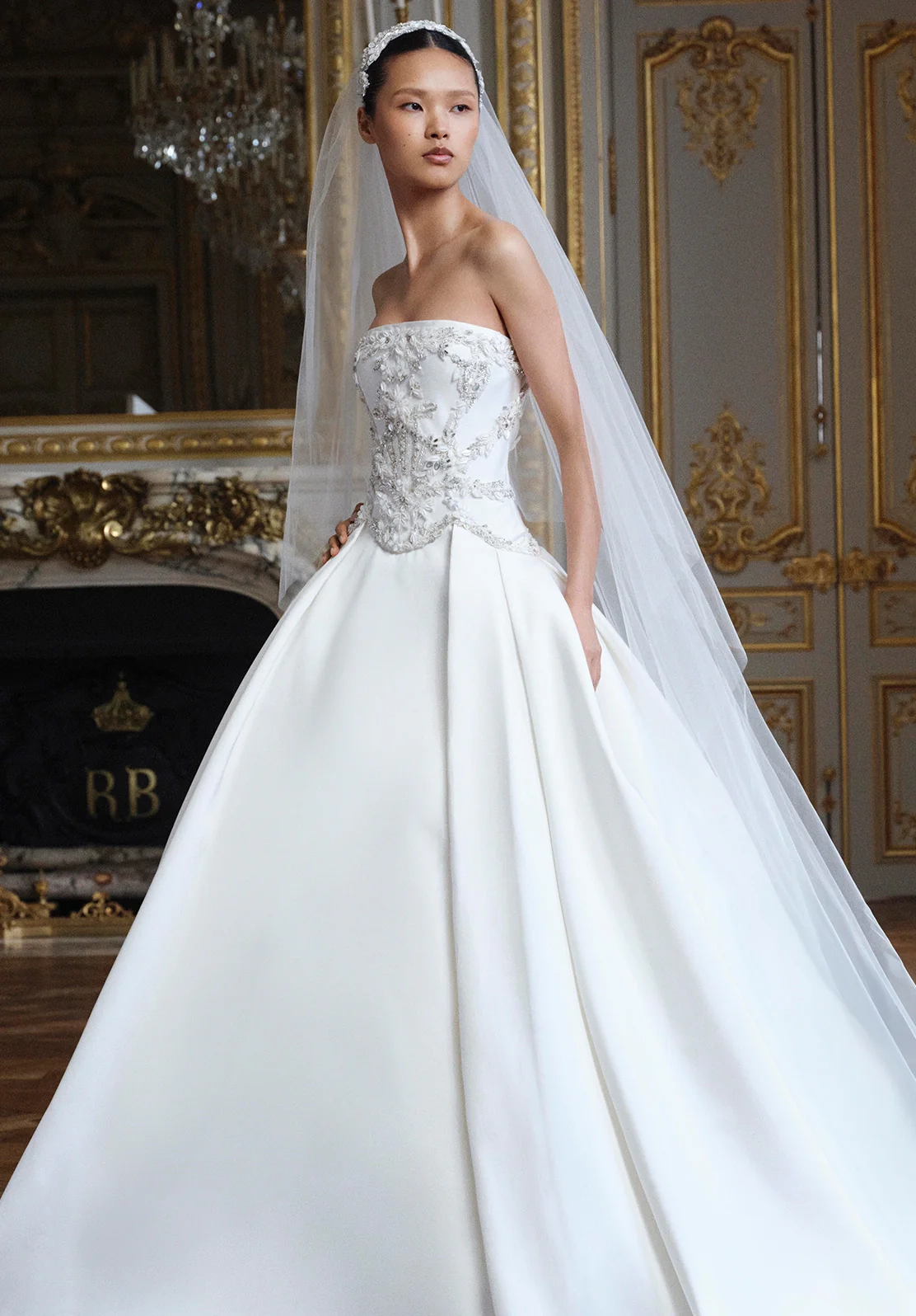 Model, lüks bir salonda, straplez, işlemeli korsajlı ve kabarık saten etekli, Elie Saab imzalı zarif bir gelinlik modeli giyerken, başındaki ışıltılı taç ve tül duvağıyla masalsı bir prenses duruşu sergiliyor.