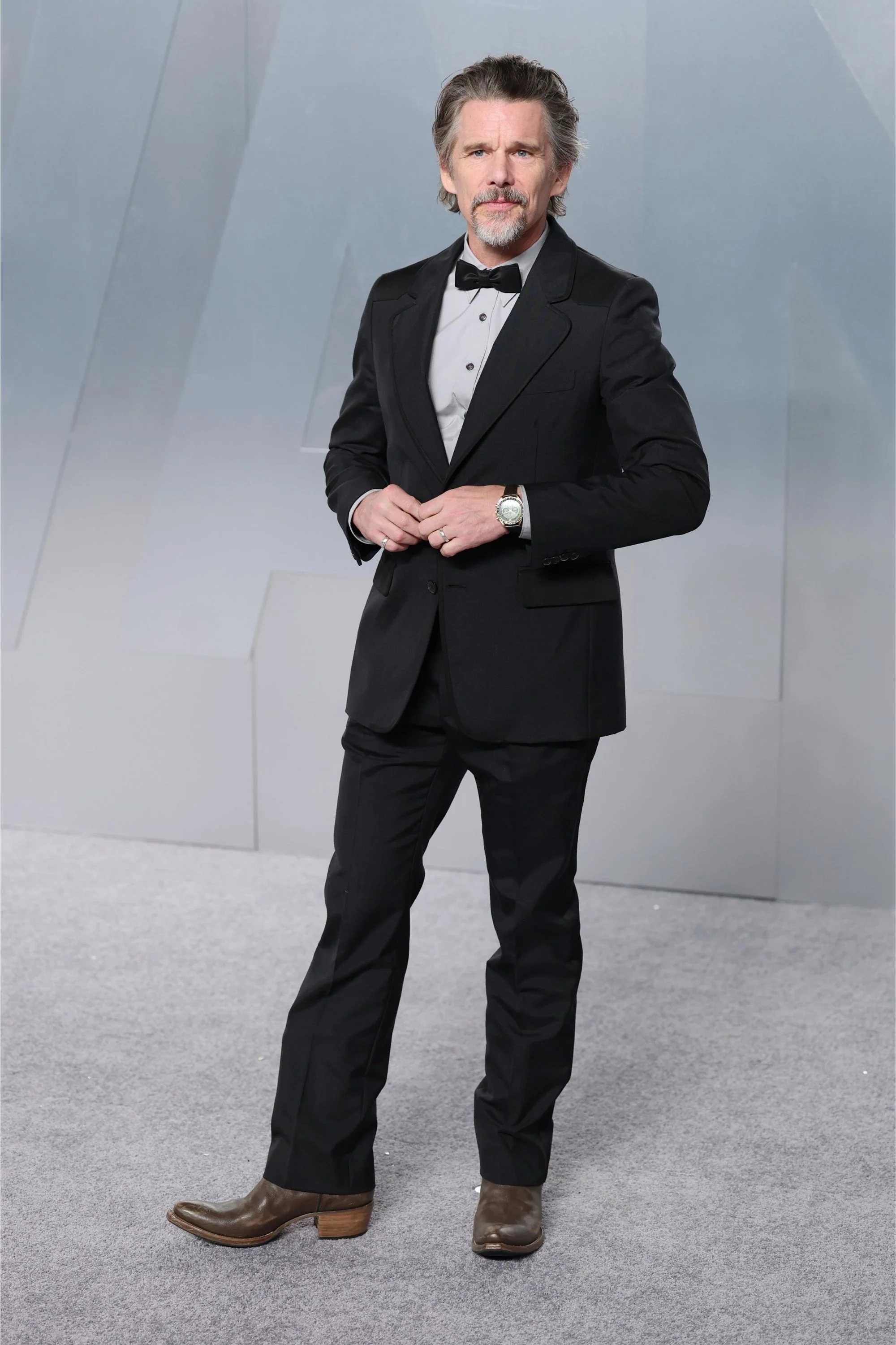 Oscar törenine katılan Ethan Hawke, siyah smokini ve kahverengi botlarıyla şık bir duruş sergilerken, sol bileğindeki altın rengi kasalı ve yeşil kadranlı özel Omega Speedmaster Moonwatch saatiyle dikkatleri üzerine çekiyor.