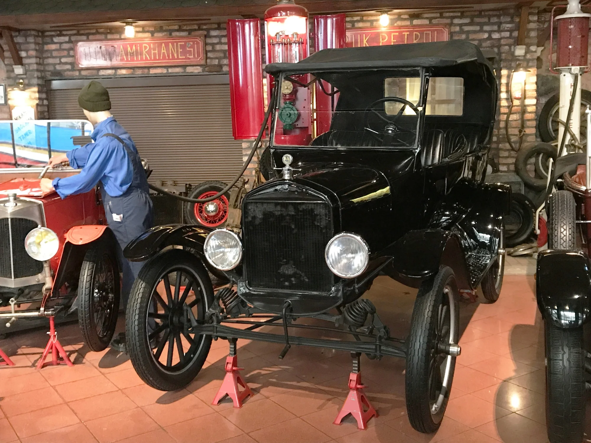 Rahmi Koç Müzesi'nde sergilenen, ön kısmı iki kırmızı kriko sehpası üzerinde yükseltilmiş, parlak siyah bir Ford Model T otomobil, tuğla duvarlı eski bir tamirhane ortamında duruyor; arka planda bir tamirci mankeni kırmızı bir araba üzerinde çalışırken, eski bir benzin pompası ve TAMİRHANESİ yazısı belirginleşiyor.