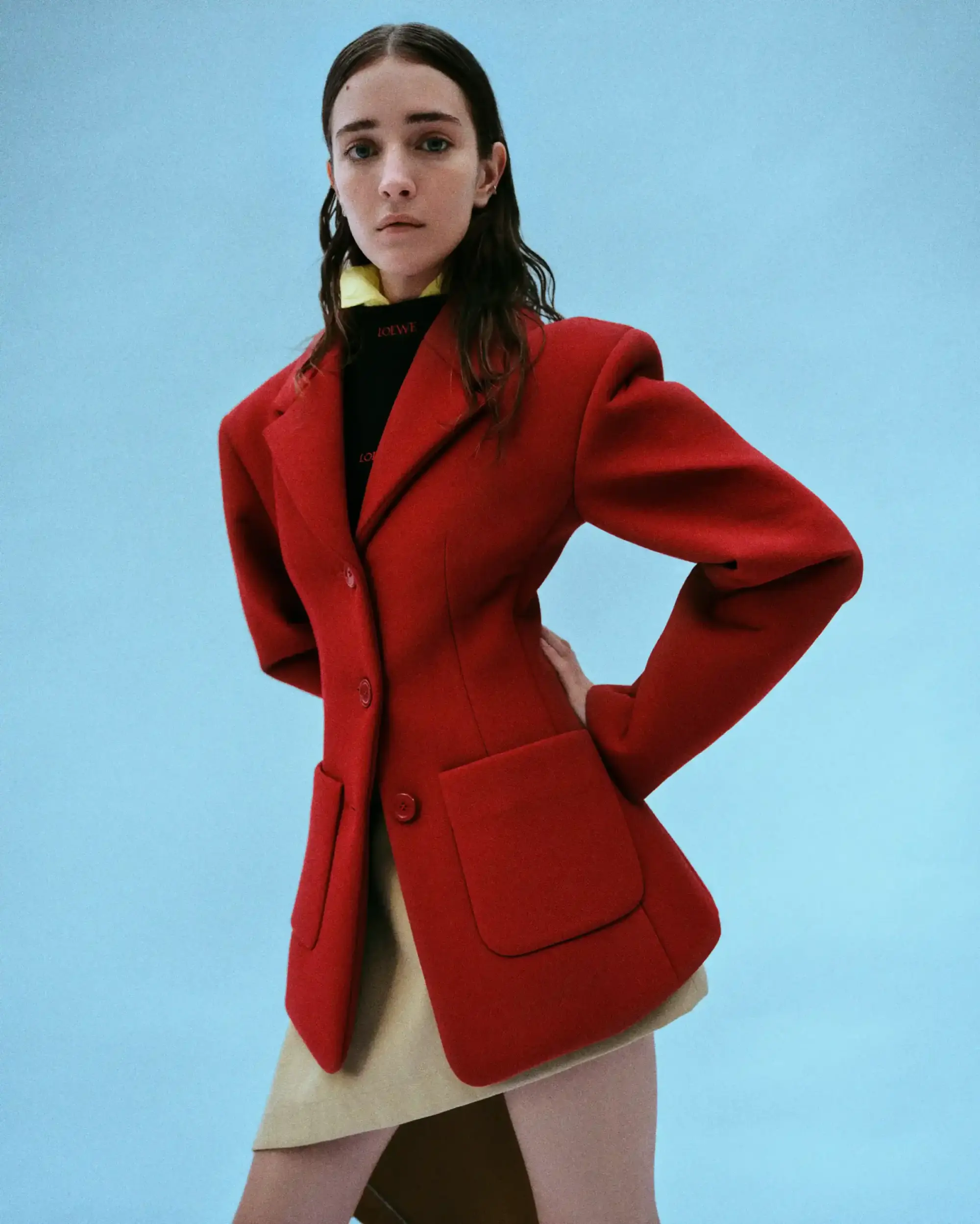 Açık mavi bir fonun önünde, güçlü omuzları olan canlı kırmızı bir blazer, siyah Loewe logolu boğazlı bir üst ve bej mini etek giyen genç bir kadın, bahar dolabına yönelik stil sahibi bir gardırop detoksu ile zamansız parçaların modern ve dinamik bir kapsül gardırop oluşturmadaki önemini yansıtıyor.
