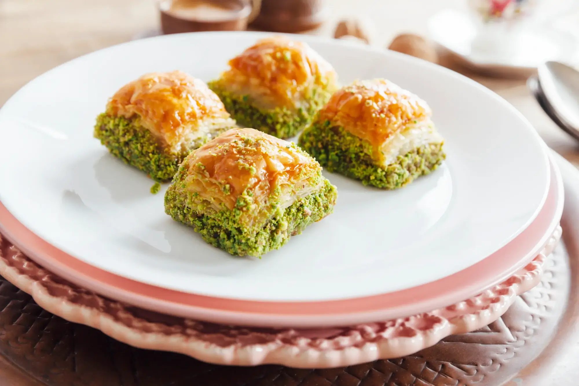 Beyaz ve pembe kenarlı tabaklarda servis edilen, üstü şerbetli ve parlak, kenarları bol yeşil fıstıkla kaplı dört adet kare fıstıklı baklava, Gaziantep'in eşsiz yemekleri arasında özel bir yere sahip bu geleneksel tatlının görselini sunuyor.