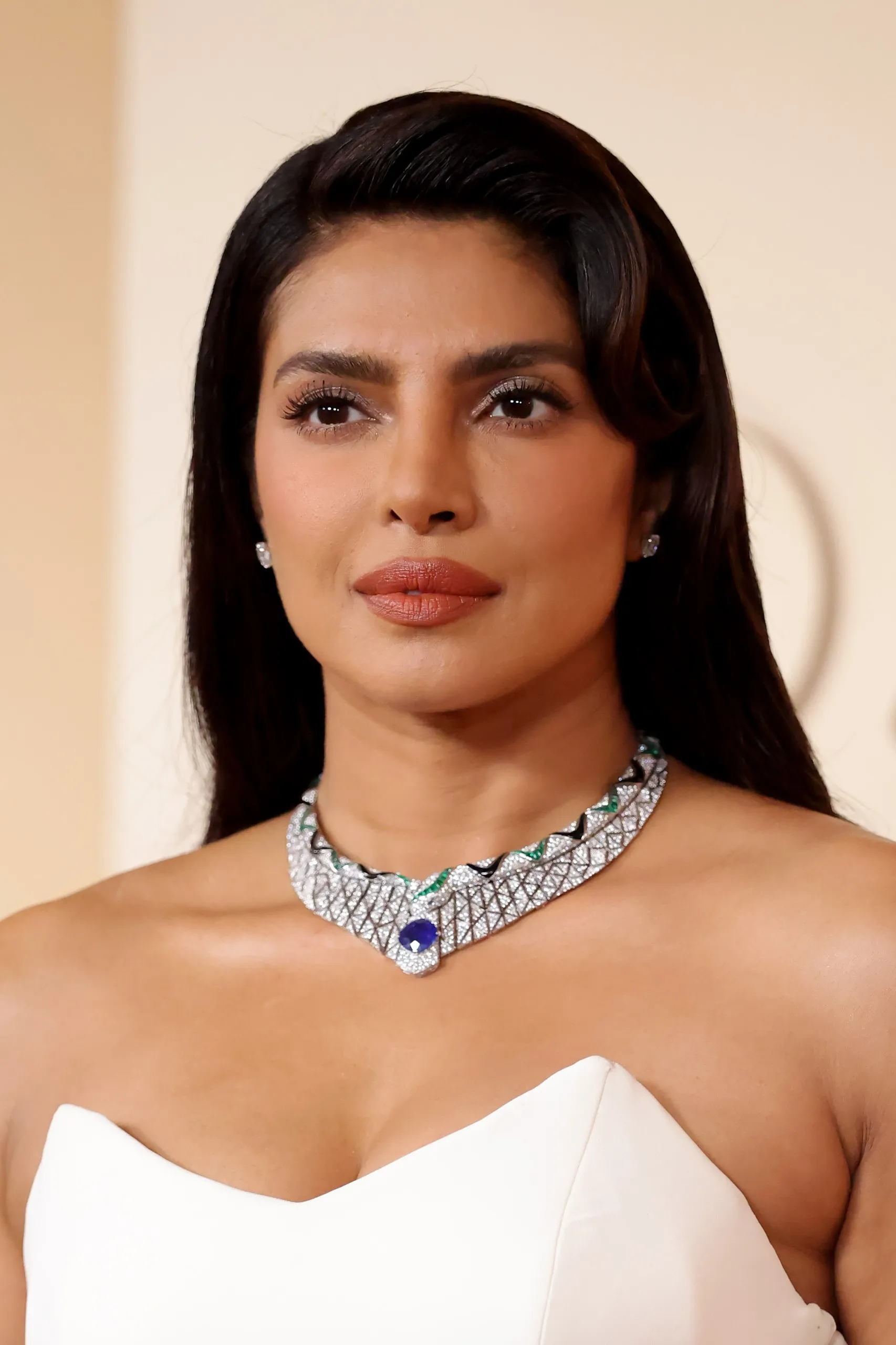 Bollywood yıldızı Priyanka Chopra Jonas'ın açık bej arka plan önünde, straplez beyaz elbisesi, zarif pırlanta küpeleri ve ortasında büyük mavi safir taşı ile zümrüt detaylı gösterişli elmas kolyesiyle doğrudan kameraya baktığı, göğüs hizasından çekilmiş yakın plan portresi.