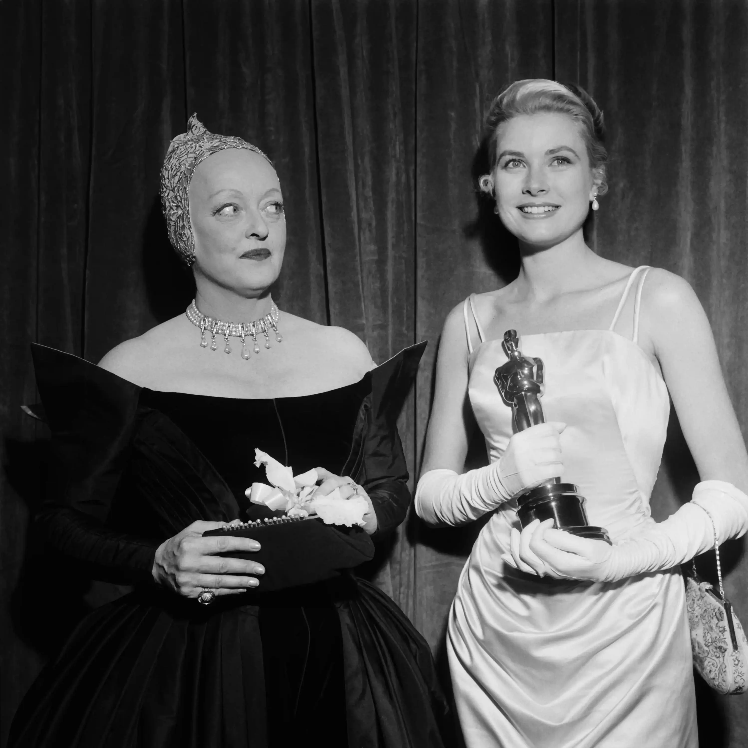 Grace Kelly’nin Oscar kazandığı bu ikonik karede, zarif Hollywood estetiği tüm sadeliğiyle öne çıkıyor. İnce askılı saten elbisesi ve uzun eldivenleriyle klasik kırmızı halı stilinin en rafine örneklerinden birini temsil eden Kelly, elinde Oscar heykelciğiyle dönemin zamansız şıklığını yansıtıyor. Yanında yer alan Bette Davis ise dramatik silueti ve heykelsi başlığıyla güçlü bir kontrast oluşturuyor; iki farklı Hollywood stil anlayışı aynı karede buluşuyor.