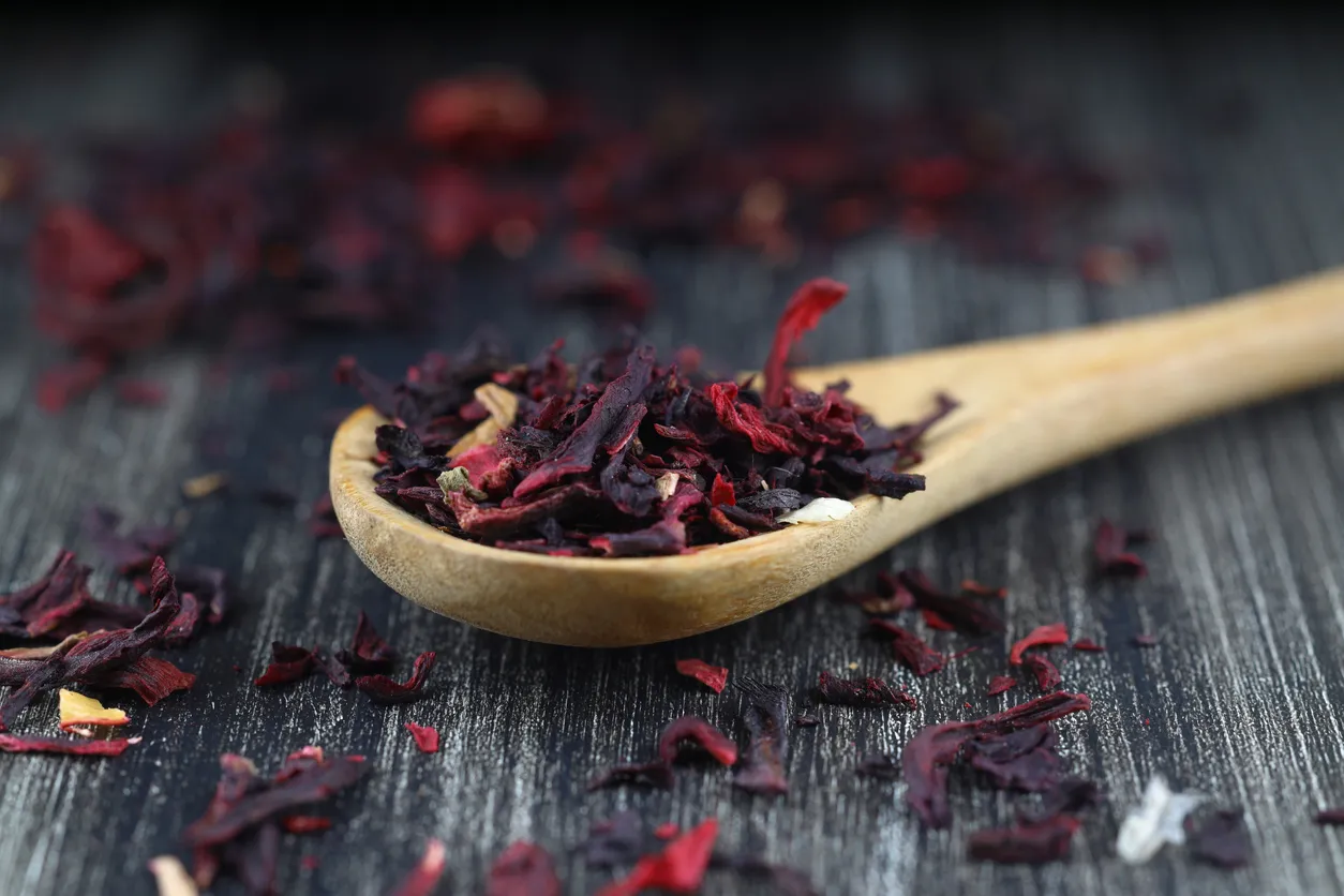 Ahşap bir kaşığın içinde, metabolizma hızlandıran ve yüksek C vitamini içeriği gibi birçok faydasıyla bilinen koyu yakut kırmızısı renginde kurutulmuş hibiskus çayı parçacıkları, çay keyfi için hazır bir şekilde duruyor.