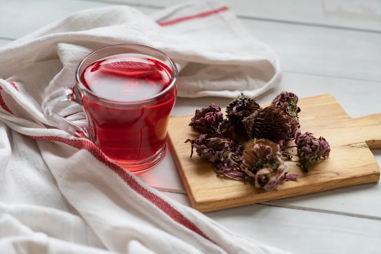 Beyaz ahşap bir zeminin üzerinde şeffaf cam bir kupada duran parlak yakut kırmızısı hibiskus çayı ve hemen yanında küçük ahşap bir kesme tahtasında demlenmeye hazır, koyu kahverengi-morumsu tonlardaki kurutulmuş hibiskus kaliksleri, bu şifa deposu bitkinin sunduğu faydaları ve metabolizma hızlandırıcı özelliklerini görselleştiriyor.