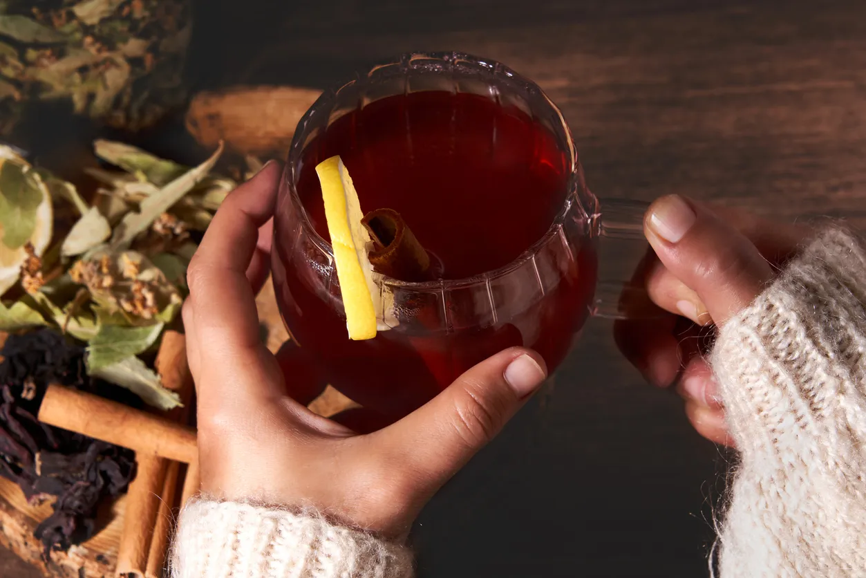 Krem rengi örgü bir kazak giymiş ellerde sıcak bir cam kupada tutulan, yakut kırmızısı rengindeki limon dilimi ve tarçın çubuğu eklenmiş hibiskus çayı, arka planda görünen kuru hibiskus çiçekleri ve tarçınlarla birlikte, içeriğindeki zengin antioksidanlar sayesinde sunduğu faydaları ve metabolizma hızlandırıcı etkilerini nazikçe hissettiriyor.