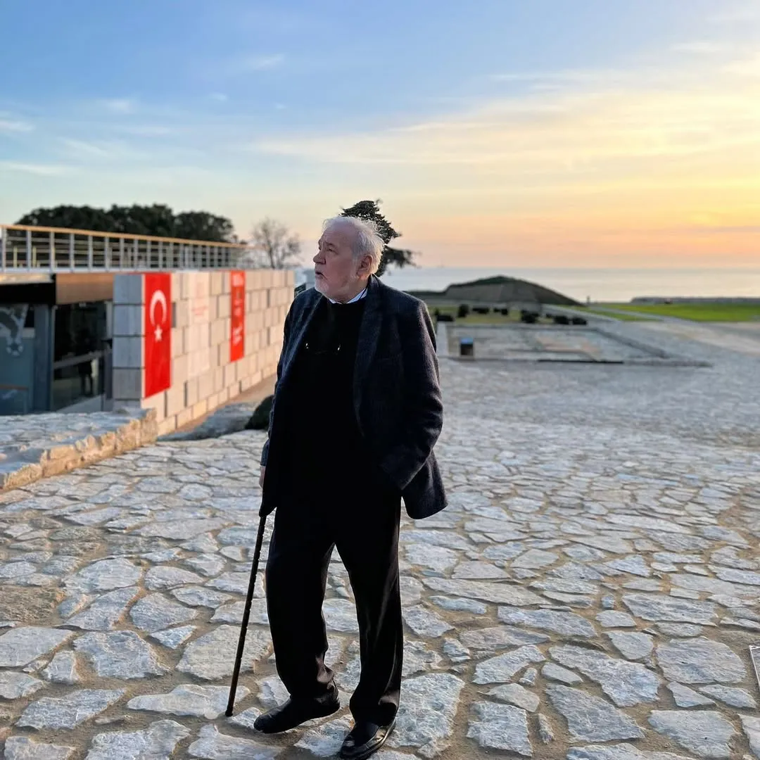 Deniz manzaralı, gün batımı kızıllığına bürünmüş gökyüzünün altında, Türk bayraklı bir binanın önündeki taş döşeli alanda bastonuyla duran ve düşünceli bir ifadeyle ufka bakarak adeta Bi Ömür Nasıl Yaşanır sorusunun derinliklerini irdeleyen Prof. Dr. İlber Ortaylı.