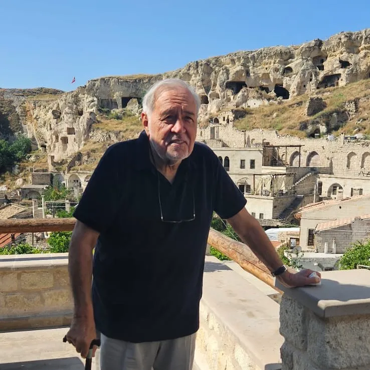 Tarihçi Ilber Ortaylı, açık mavi gökyüzü altında, Türkiye'nin Kapadokya bölgesindeki ikonik kaya oluşumları ve oyma mağara evleriyle dolu antik yerleşim yerinin önünde, bastonuna yaslanarak duruyor, üzerinde gözlüğü asılı lacivert tişörtüyle dikkat çekiyor.