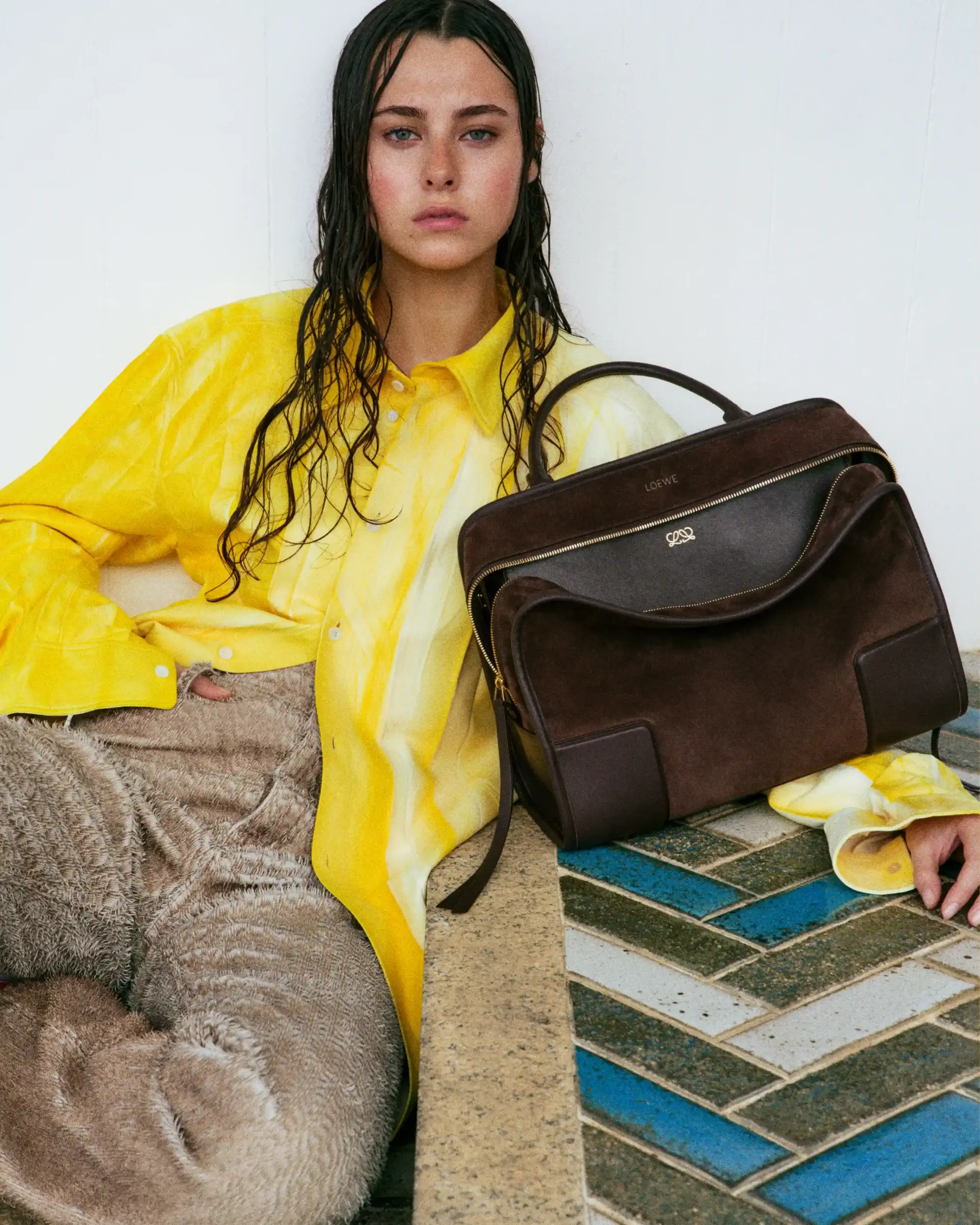Genç bir kadın, sarı batik desenli gömleği, dokulu bej pantolonu ve ıslak saçlarıyla, yanında duran Loewe çantası eşliğinde, ilkbahar-yaz kapsül gardıroplar için renk ve desen dengesini yansıtan modern bir stil sunuyor.