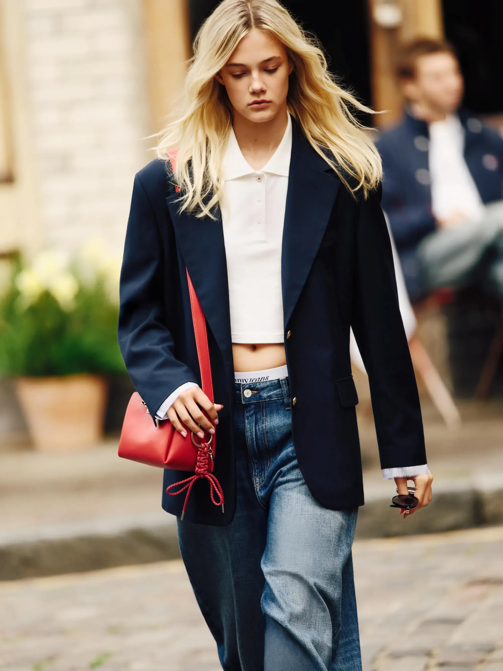 Genç, sarışın bir kadın, oversize lacivert bir blazer ceket, beyaz kısa kollu polo yaka bir tişört ve açık mavi düz kesim jean giyerek, kırmızı minimal çantasıyla bir ilkbahar-yaz kapsül gardırop için temel parçaları şık bir şekilde birleştiriyor.