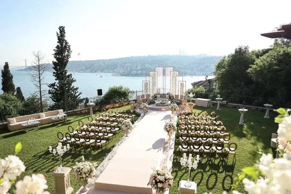 Boğaz'ın büyüleyici manzarasına karşı, yemyeşil bir bahçede, beyaz çiçeklerle bezenmiş zarif bir düğün veya açık hava kutlama alanı; altın detaylı sandalyeler, beyaz halı kaplı bir yürüyüş yolu ve modern mimarili sahne arkası dekorasyonuyla özel bir gün için hazırlanmış.