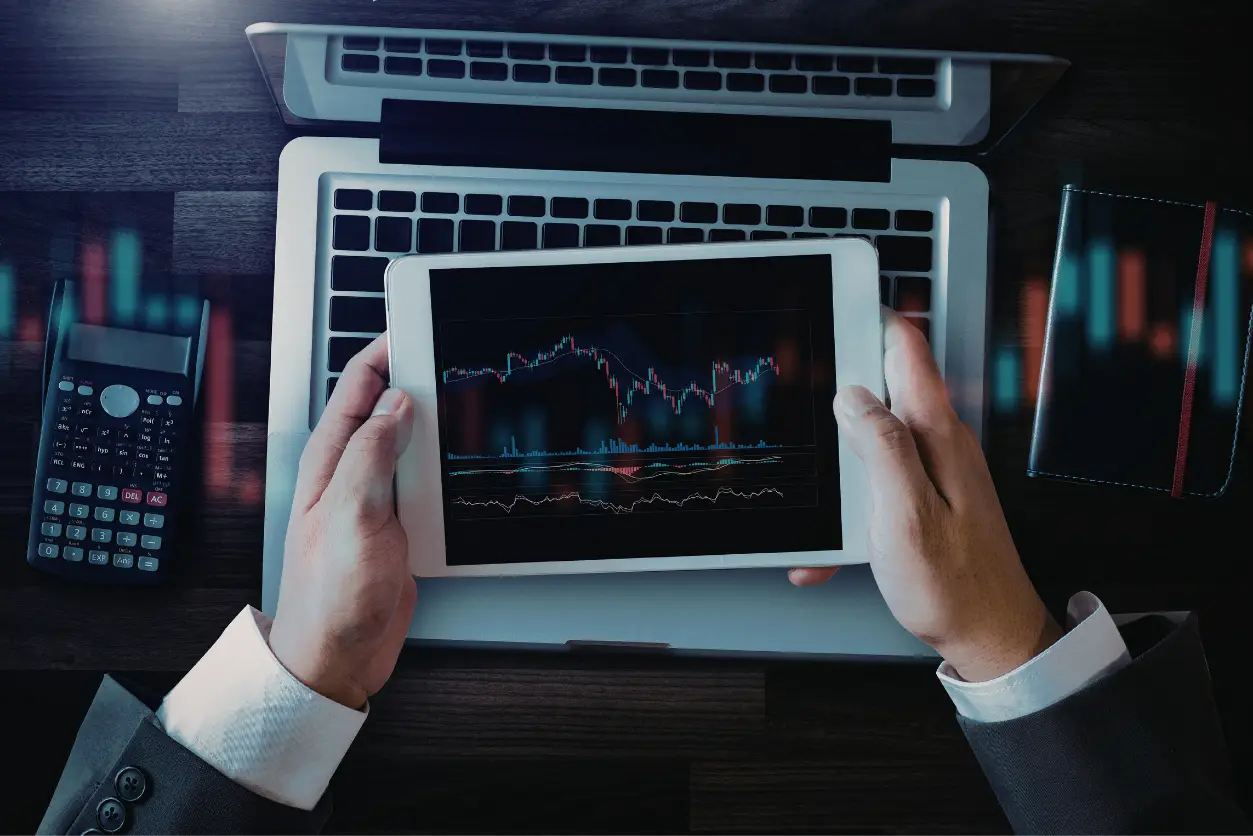 Bir iş profesyonelinin dizüstü bilgisayarın üzerinde finansal grafikler gösteren bir tableti iki eliyle tutarak, yanında bir hesap makinesi ve not defteriyle karmaşık piyasa verilerini dikkatle incelediği bu görsel, uzun süreli analiz ve ardışık kararların getirebileceği karar yorgunluğunu çağrıştırıyor.