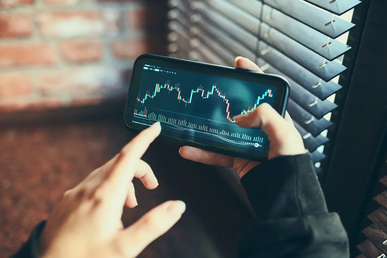 Finansal piyasalardaki karmaşık veri grafiklerini gösteren bir akıllı telefonu parmaklarıyla aktif olarak inceleyen bir kişi, sürekli veri akışıyla ortaya çıkan karar yorgunluğunun potansiyel etkilerini yansıtıyor.