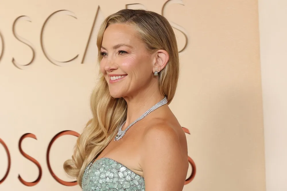 Sarışın, uzun dalgalı saçları ve ışıltılı pırlanta kolyesiyle zarif bir şekilde gülümseyen Kate Hudson, gümüş payetli açık mavi straplez elbisesi ve aynı tarzda pırlanta küpeleriyle şık duruşuyla kırmızı halıda göz kamaştırıyor, arka planda belirgin Oscar yazısı görülüyor.