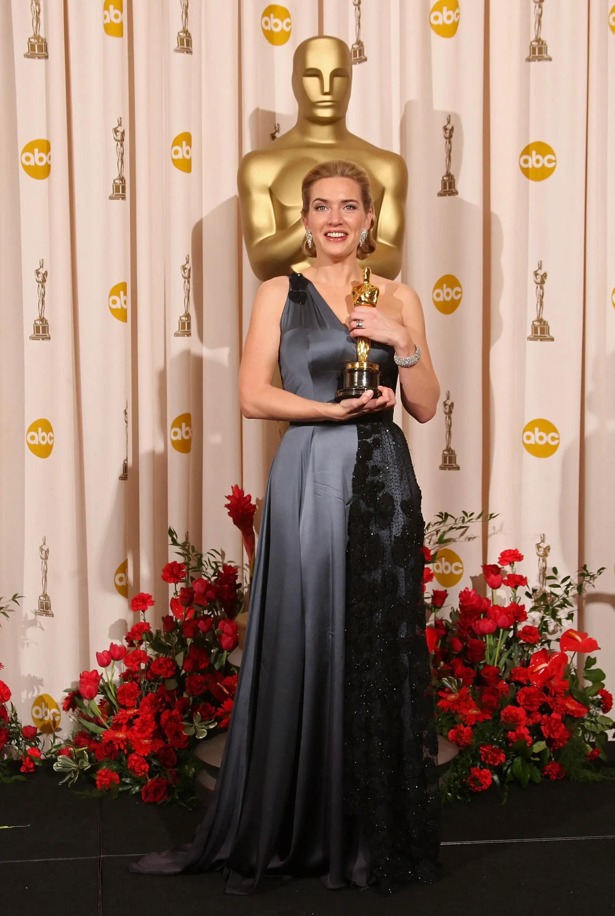 Kate Winslet, bu karede Oscar heykelciğini elinde tutarken klasik Hollywood zarafetini çağdaş bir dokunuşla yorumluyor. Tek omuzlu, gri tonlardaki saten elbise; siyah işlemeli panel detayıyla dramatik bir kontrast yaratırken sade ama güçlü bir siluet oluşturuyor. Minimal mücevher tercihleri ve geriye doğru toplanmış saçları görünümün sofistike karakterini destekliyor. Sonuçta ortaya çıkan stil, abartıya kaçmadan kırmızı halıda güçlü bir etki bırakmanın en iyi örneklerinden biri.