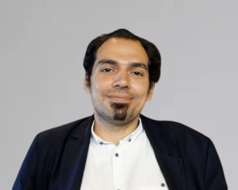 Kerem Akça, gri düz bir arka plan önünde, koyu renk saçları geriye taranmış, hafifçe gülümseyen ve çene sakalı olan bir beyefendi olarak beyaz düğmeli gömleği ve şık lacivert ceketiyle kameraya odaklanmış bir şekilde bakıyor.