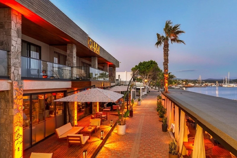 Bodrum'daki Khai Hotels'in akşam ışıklarıyla aydınlatılmış modern taş ve ahşap mimariye sahip binası, geniş ahşap terasında rahat oturma alanları ve şemsiyelerle donatılmış davetkar bir dış mekan lounge sunarken, hemen yanında palmiye ağaçları ve ışıklı sahil yolu boyunca uzanan yelkenli teknelerle dolu marinayı gösteren büyüleyici bir sahil manzarası bulunmaktadır.