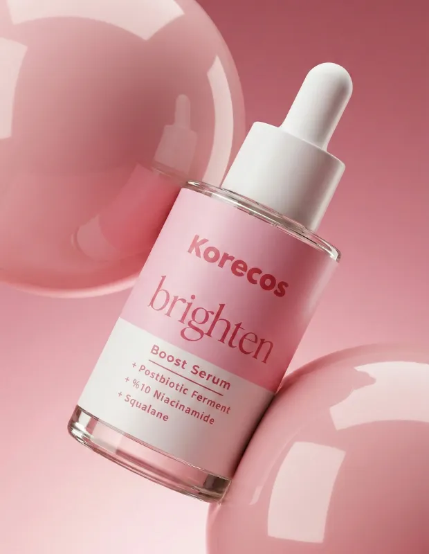 Pembe etiketli ve beyaz damlalıklı kapağı olan Korecos brighten Boost Serum şişesi, Postbiotic Ferment, Niasinamid ve Squalane içeren formülüyle cilde yönelik yeni ürün ve gelişmeler arasında dikkat çeken parlak pembe kürelerin arasında eğik duruyor.
