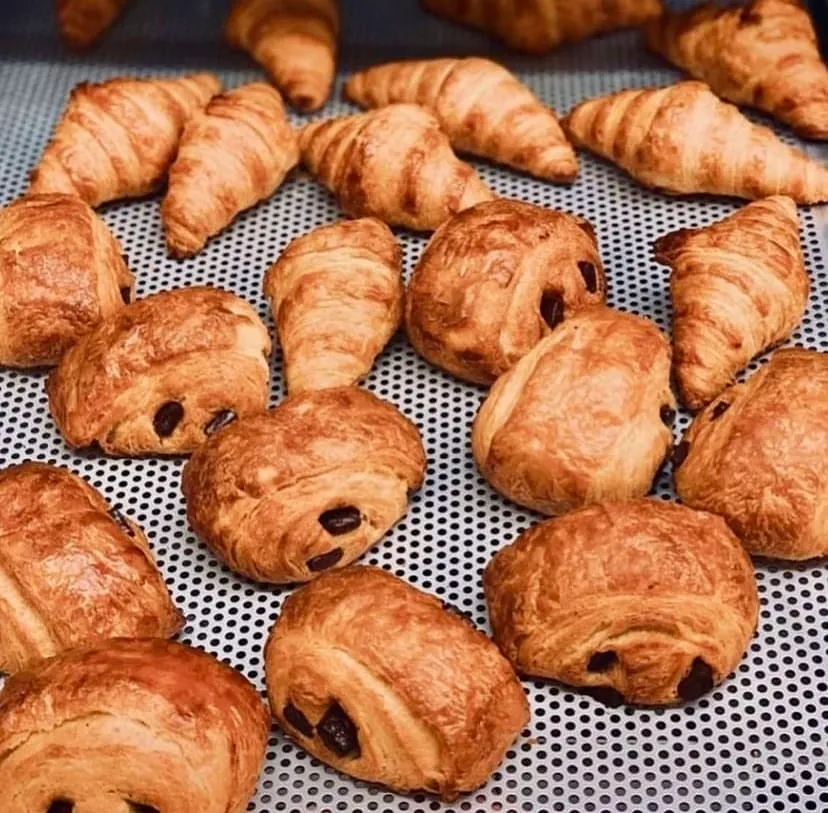 Metal bir tepsi üzerinde, taptaze pişmiş, nar gibi kızarmış ve kat kat görünen çok sayıda klasik kruvasan ile erimiş çikolata parçacıklarıyla dolu pain au chocolat hamur işleri iştah açıcı bir şekilde sergileniyor.