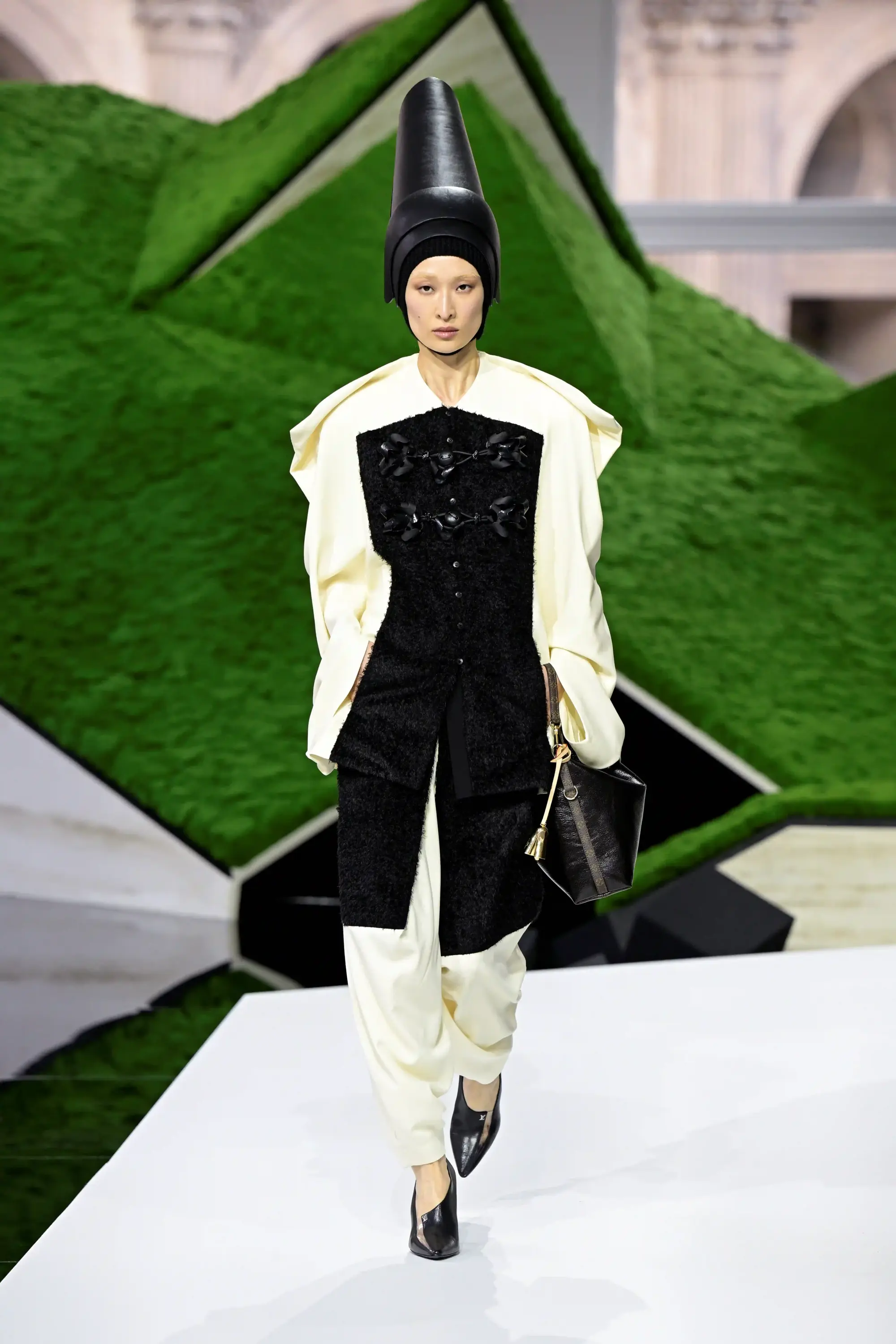 Model, Louis Vuitton Sonbahar-Kış 2026 koleksiyonundan fütüristik bir neo-manzara podyumunda, sivri siyah bir şapka, kabarık bej bir üst, siyah tüylü, dekoratif bağlamalı bir yelek ve bej bol paça pantolon giymiş, elinde siyah Louis Vuitton çantasıyla yürürken, doğa ve teknolojiyi birleştiren tasarımların sergilendiği moda haftasının özgün atmosferini yansıtıyor.