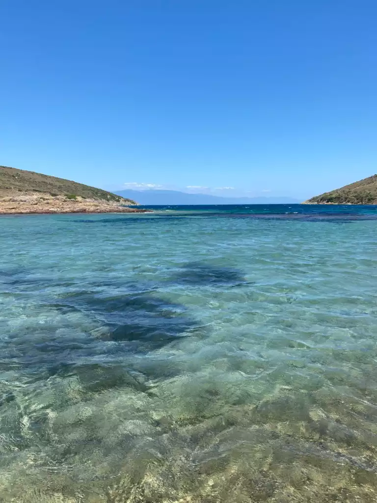 Cunda yakınlarındaki Maden Adası'nın berrak turkuaz sularının ortasında, sol tarafta kayalık bir koy, sağ tarafta yeşil tepelerle çevrili, güneşli bir günde uzaktan dağların seçildiği sakin ve doğal bir deniz manzarası.