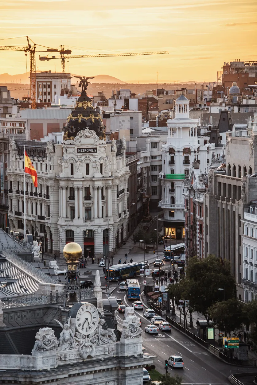 Madrid'in altın rengi gün batımında çekilmiş büyüleyici şehir manzarası; tepesinde heykel bulunan ikonik Metrópolis Binası'nı, İspanya bayrağını, zarif saat kulesini ve hareketli caddelerdeki araçlarla insanları gözler önüne sererek eşsiz bir seyahat atmosferi sunuyor.