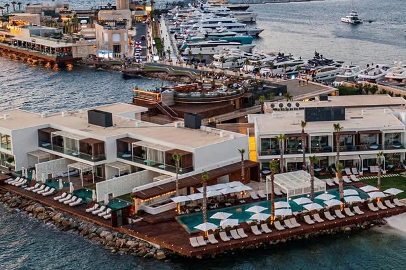 Marinaya demirlemiş çok sayıda lüks yatın bulunduğu bir koyda yer alan, modern mimariye sahip beyaz binaları, çok katlı odaları, denize nazır havuzları ve şezlonglarıyla bir marina otelin kuşbakışı, ışıltılı akşam görüntüsü.