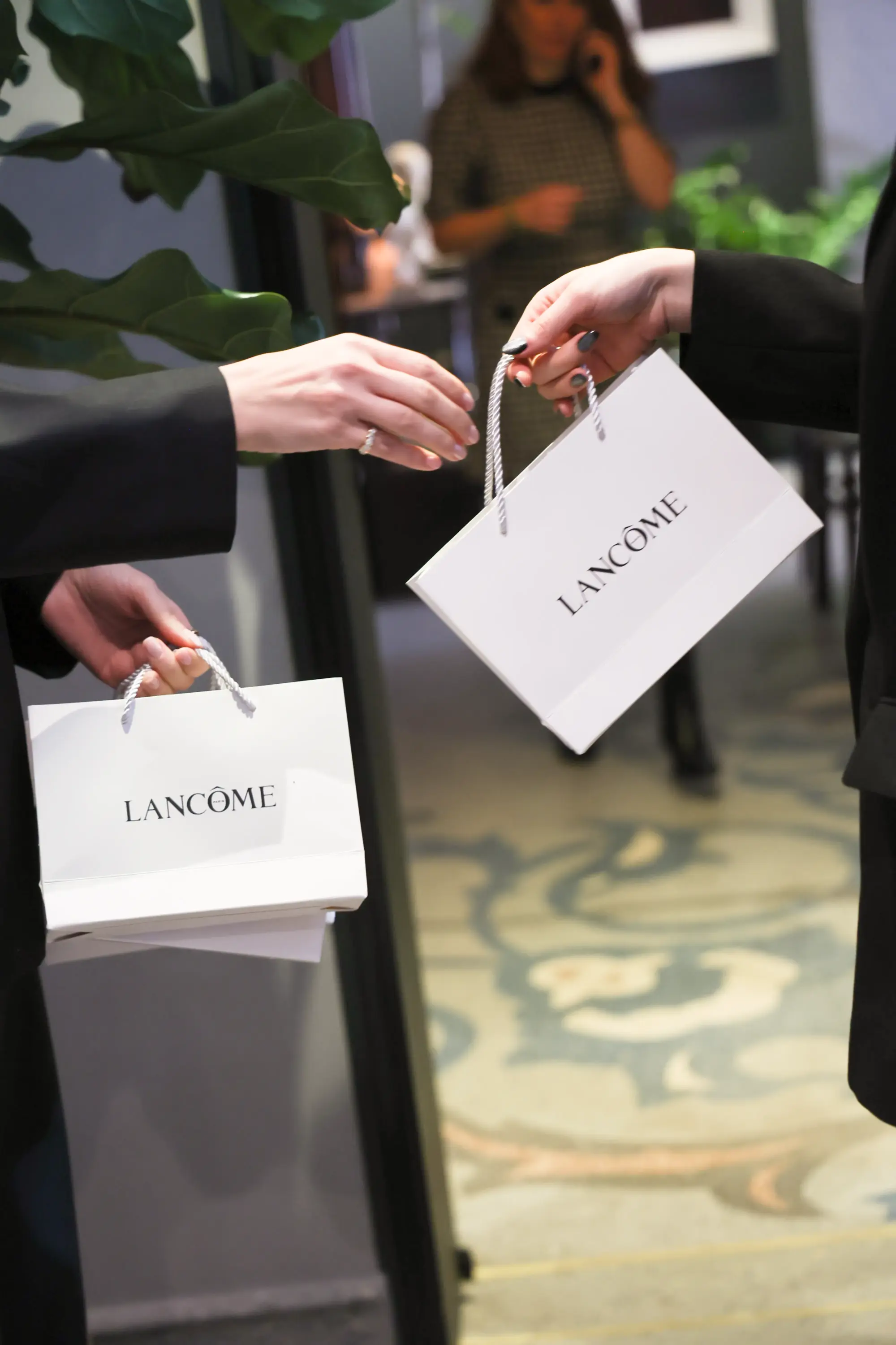 Şık bir etkinlik veya davet ortamında, siyah ceketli iki kişinin özenli elleri arasında, üzerinde Lancôme markasının zarif logosu bulunan beyaz hediye çantaları nazikçe el değiştiriyor, bu an lüks kozmetik ve kişisel bakım ürünlerine yönelik özel bir deneyimin başlangıcını yansıtıyor.