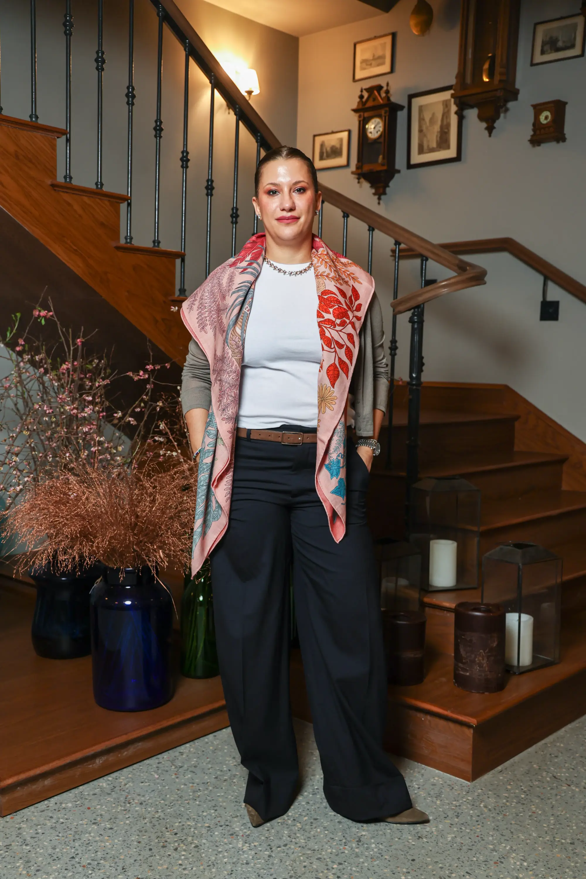 Hovagimyan, Oggusto etkinliğinde, saçları sıkıca toplanmış ve hafif makyajlı bir şekilde, beyaz tişörtü, açık kahve hırkası ve lacivert geniş paça pantolonu üzerine pembe tonlarında yaprak desenli büyük bir şal takarak, antika saatlerin asılı olduğu ahşap bir merdivenin önünde duruyor.