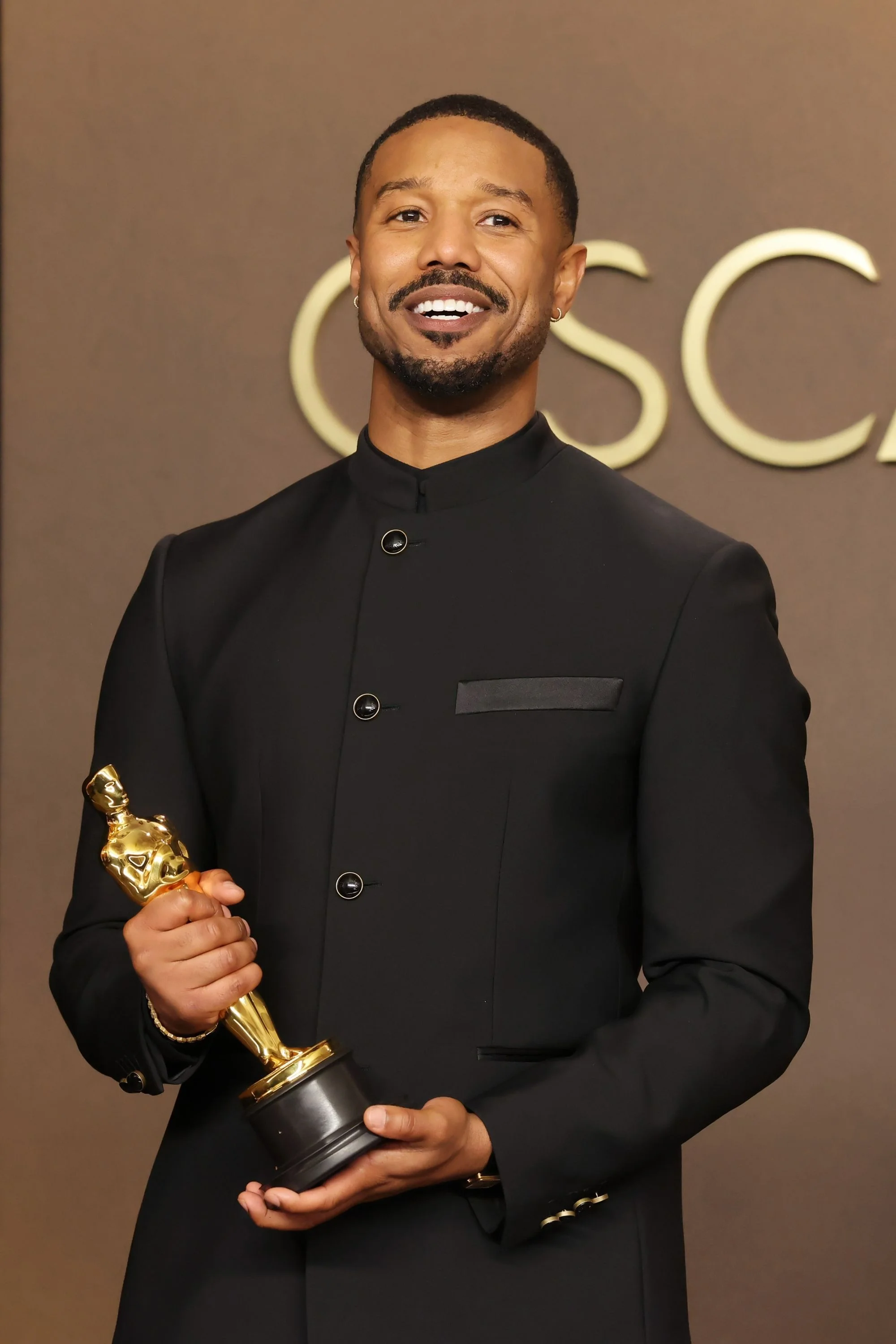 Michael B. Jordan, siyah hakim yaka bir ceket giymiş, gülümseyerek doğrudan kameraya bakıyor, parlayan altın rengi bir Oscar ödülünü iki eliyle tutuyor ve arkasında kısmen görünen büyük altın harflerle yazılı Oscar logosu yer alıyor.