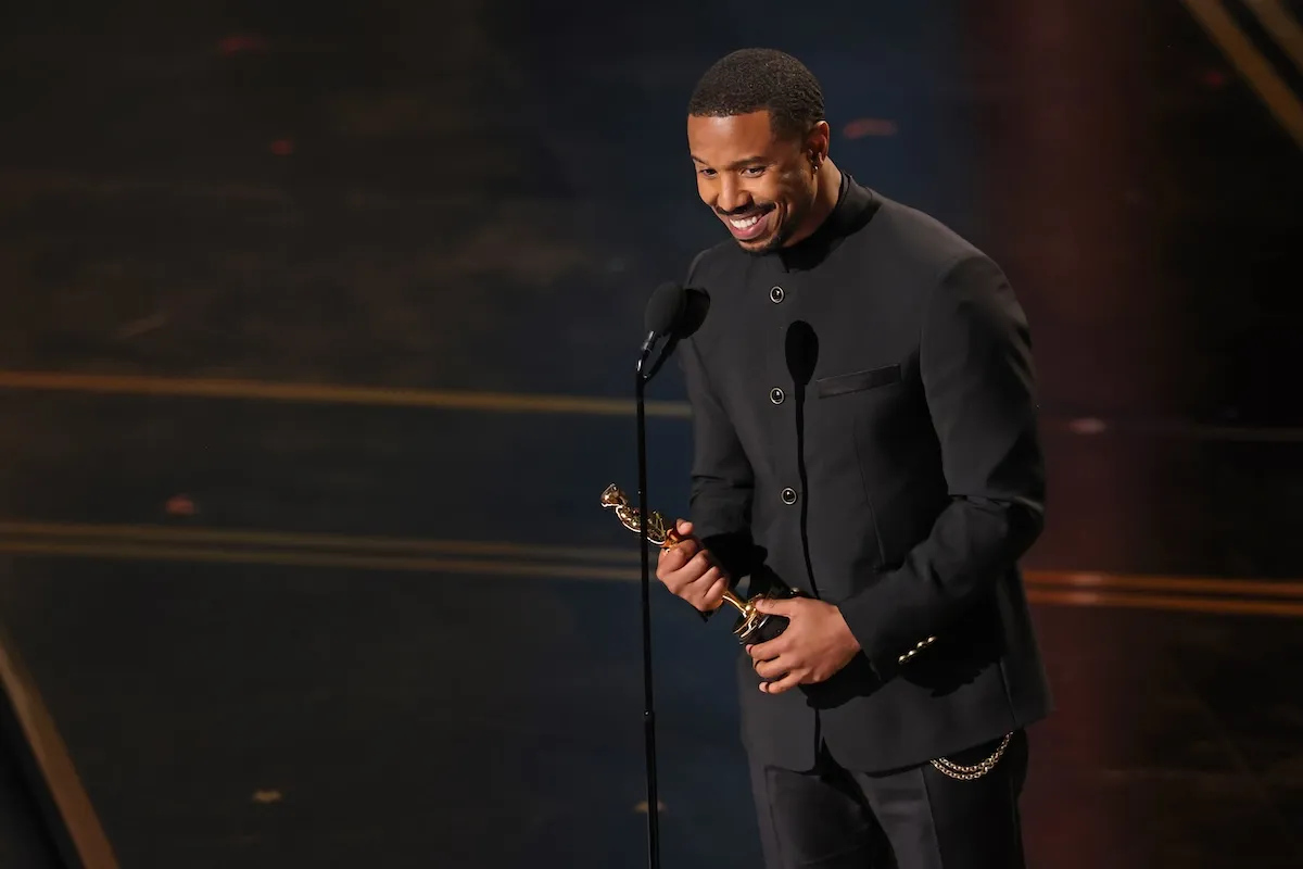 Michael B. Jordan, 2026 Akademi Ödülleri'nde En İyi Erkek Oyuncu Oscar Ödülü'nü kucaklarken, siyah dik yakalı takım elbisesiyle sahnedeki mikrofonun önünde içten bir gülümsemeyle duruyor.