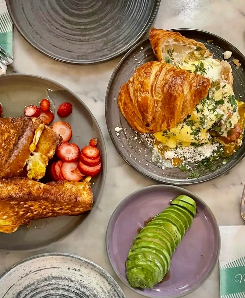 Mums Cafe'de sunulan zengin bir kahvaltı masası görüntüsü; çırpılmış yumurta, peynir ve taze yeşilliklerle doldurulmuş altın rengi, çıtır çıtır bir kruvasan, yanında çilek dilimleriyle ballı Fransız tostu ve dilimlenmiş avokado ile doludur.