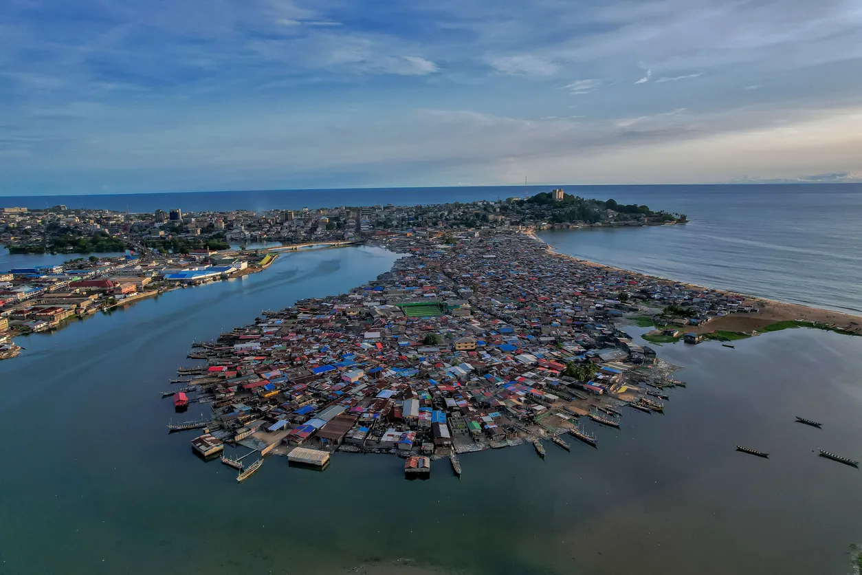 Monrovia'nın yoğun nüfuslu, renkli evlerle dolu kıyı yerleşiminin kuşbakışı görüntüsü, bir tarafta Atlantik Okyanusu, diğer tarafta nehir veya lagün suları arasında uzanan dar bir yarımadayı gösterirken, köprüler ve şehir merkezinin silüeti ufukta beliriyor.
