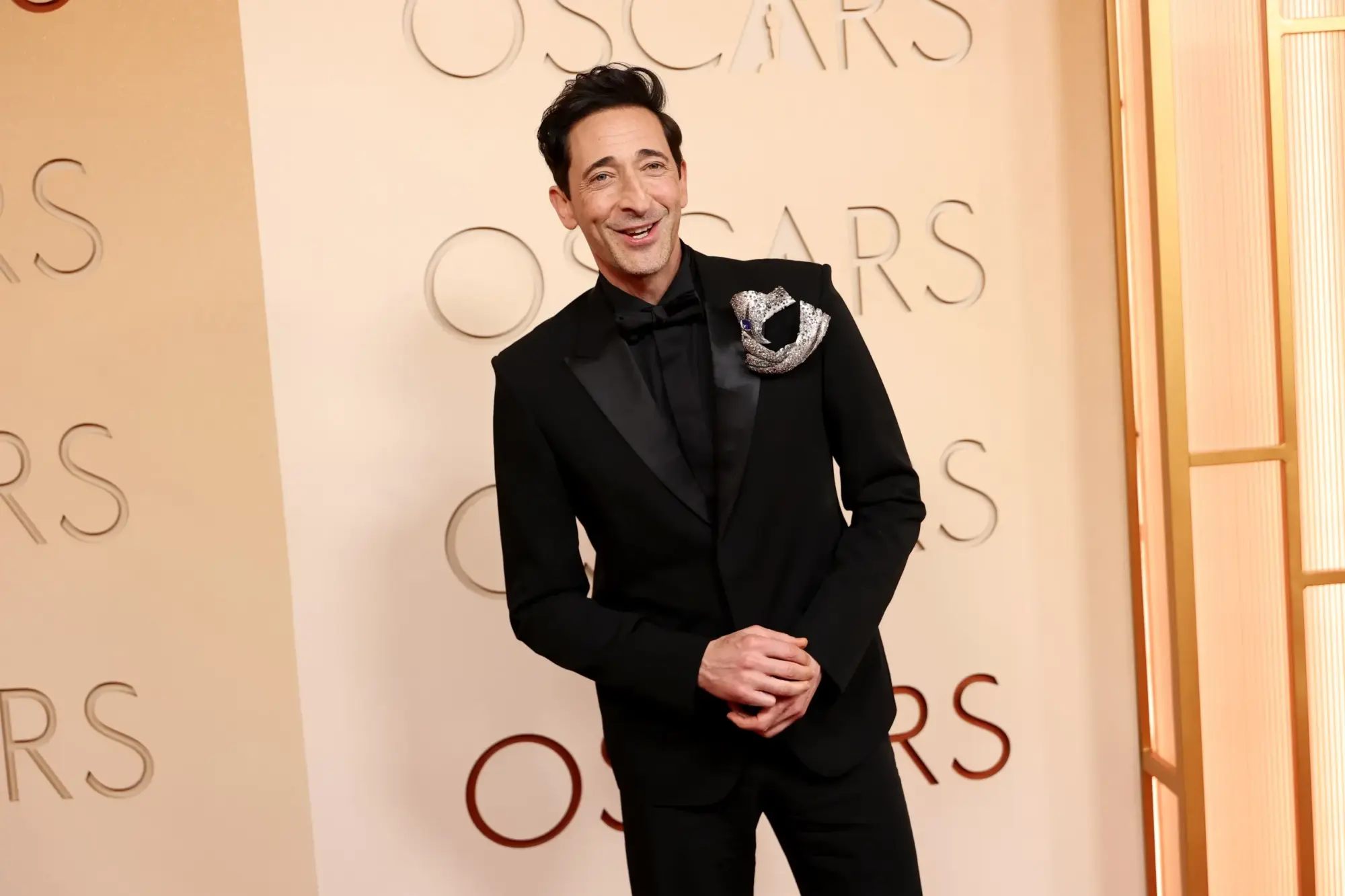 Aktör Adrien Brody, arkasında tekrar eden OSCARS yazılarının ve sağda dikey altın panellerin bulunduğu açık renkli bir fonda, siyah smokini ve yakasındaki göz alıcı gümüş broşuyla kameraya doğru içtenlikle gülümseyerek ellerini kavuşturmuş şekilde duruyor.