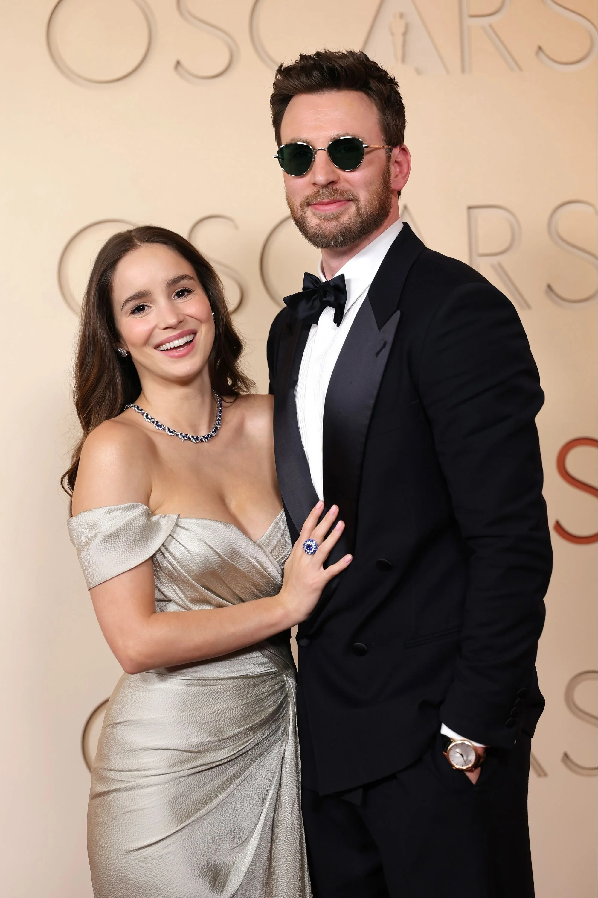 Oscar etkinliğinde eşi Alba Baptista ile birlikte gülümseyerek poz veren Chris Evans, siyah takım elbisesi, güneş gözlüğü ve bileğindeki ışığı yansıtan gold-toned Chopard L.U.C kol saatiyle dikkat çekerken, Baptista parlak gümüş rengi straplez elbisesi ve safir detaylı mücevherleriyle zarif bir duruş sergiliyor.