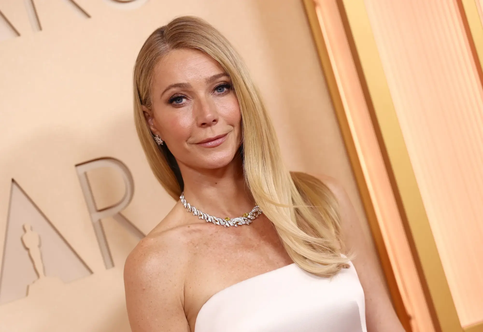 Gwyneth Paltrow, uzun sarı saçları omuzlarından aşağı süzülürken, omuzlarını açıkta bırakan beyaz straplez elbisesi ve parıldayan pırlanta takılarıyla, Oscar yazısının görüldüğü bir etkinlikte kameraya hafifçe gülümseyerek poz veriyor.